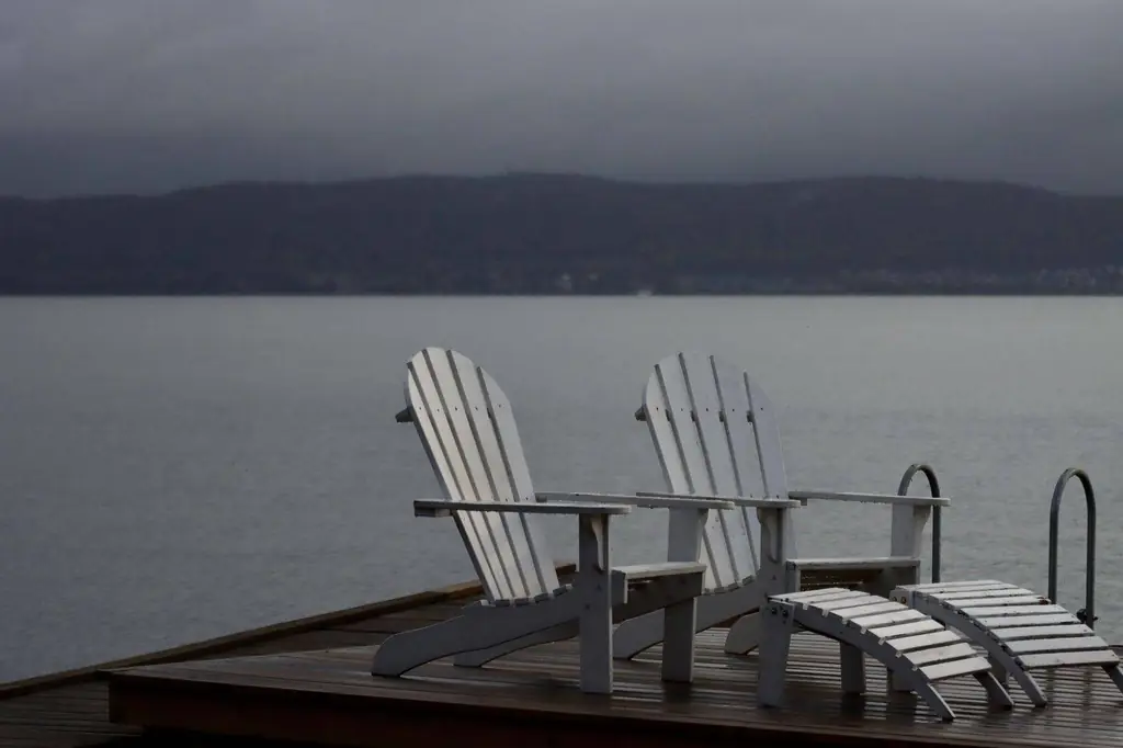 Two Adirondack chairs on a wooden dock by a serene lake under cloudy skies