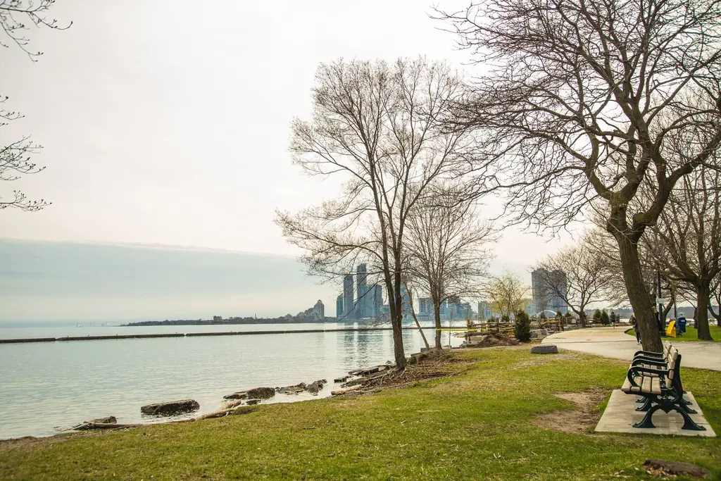 Scenery of Detroit skyline from peaceful Windsor Riverfront Park with green lawns and leafless trees against cloudy sunset