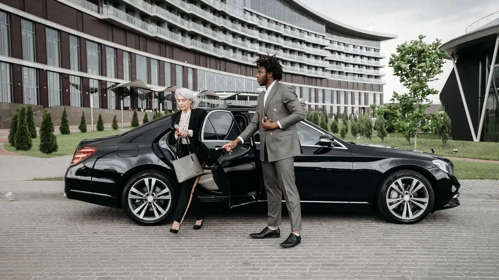 A professional chauffeur assists a businesswoman exiting a luxury car in front of a modern building