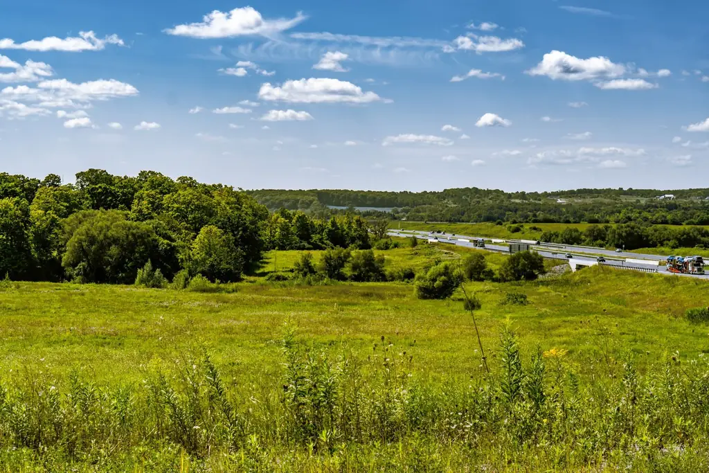 A wide view of a highway cutting through lush meadows and fields under a bright blue sky in Pickering, Canada