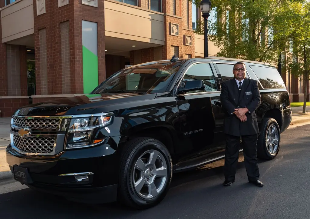 A professional chauffeur stands next to a black luxury SUV outside a modern building in daylight