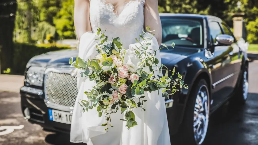 Bride in white gown holding a bouquet near a luxury car, symbolizing elegance and romance